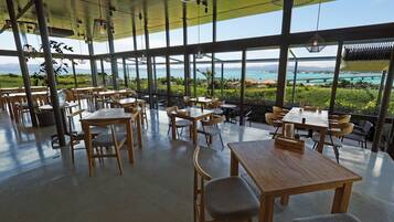 Un vaste espace intérieur aménagé en salle à manger, doté de grandes fenêtres offrant une vue sur un paysage côtier. Des tables et des chaises sont disposées pour les repas.