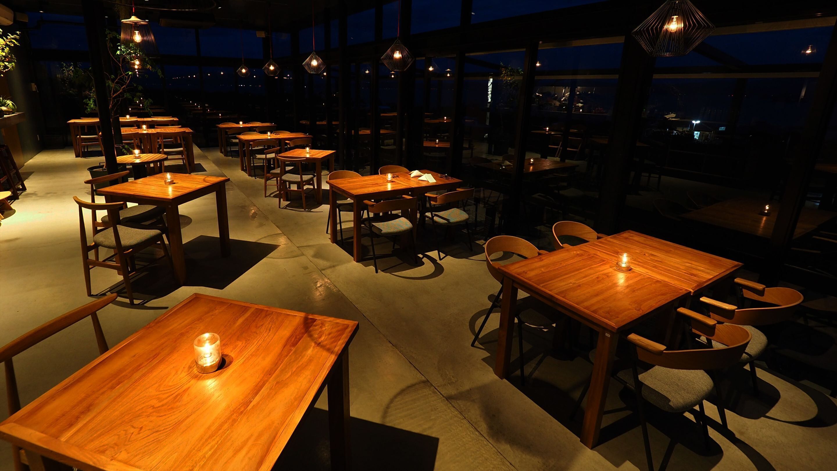Un restaurante con mesas y sillas de madera, ventanas amplias y lámparas colgantes.