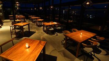 Un restaurante con mesas y sillas de madera, ventanas amplias y lámparas colgantes.