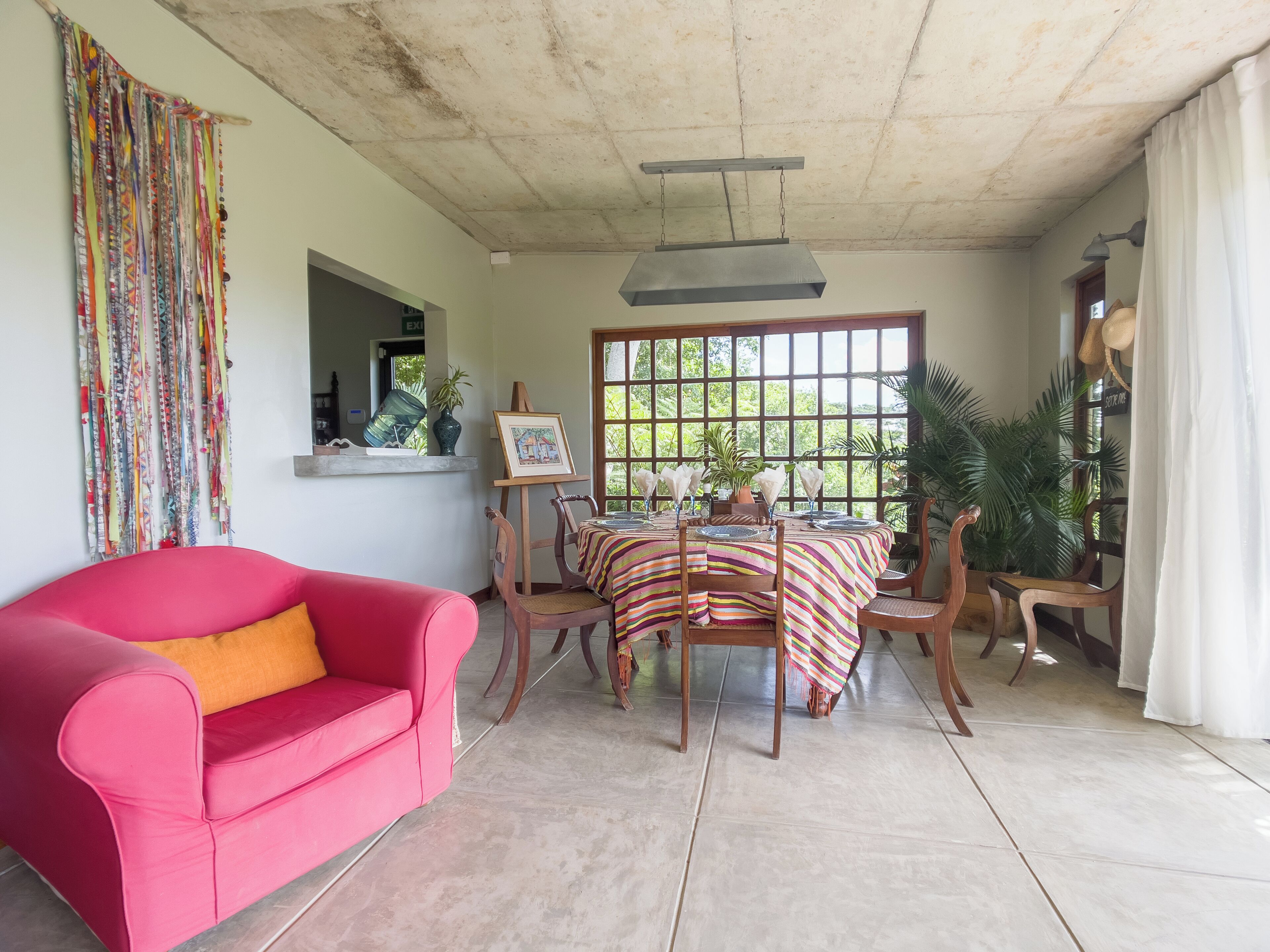 Un coin repas avec une table dressée pour quatre personnes, entourée de chaises, d’un canapé rouge et d’une fenêtre donnant sur un paysage verdoyant.