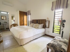 A bedroom with a large bed, a wooden wardrobe, a bedside table, a chair, and a window with a view of greenery. - Green Cottage Chamarel (Chamarel)