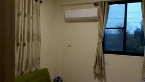 Una habitación con sofá verde, una mesita redonda con un florero, una ventana con cortinas y un equipo de aire acondicionado.