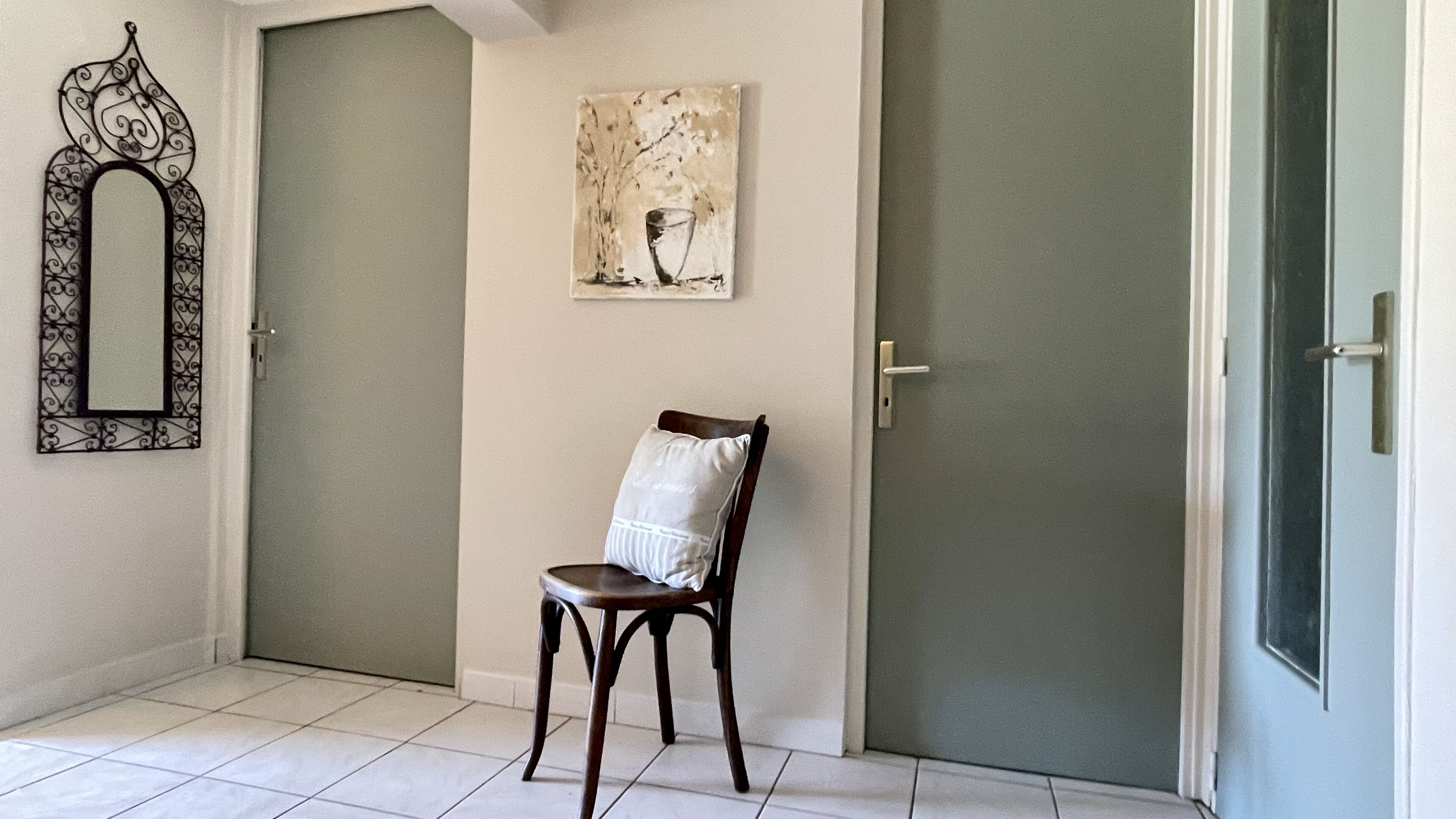 Un pasillo con dos puertas cerradas, una silla con cojín, un espejo en la pared y un cuadro.
