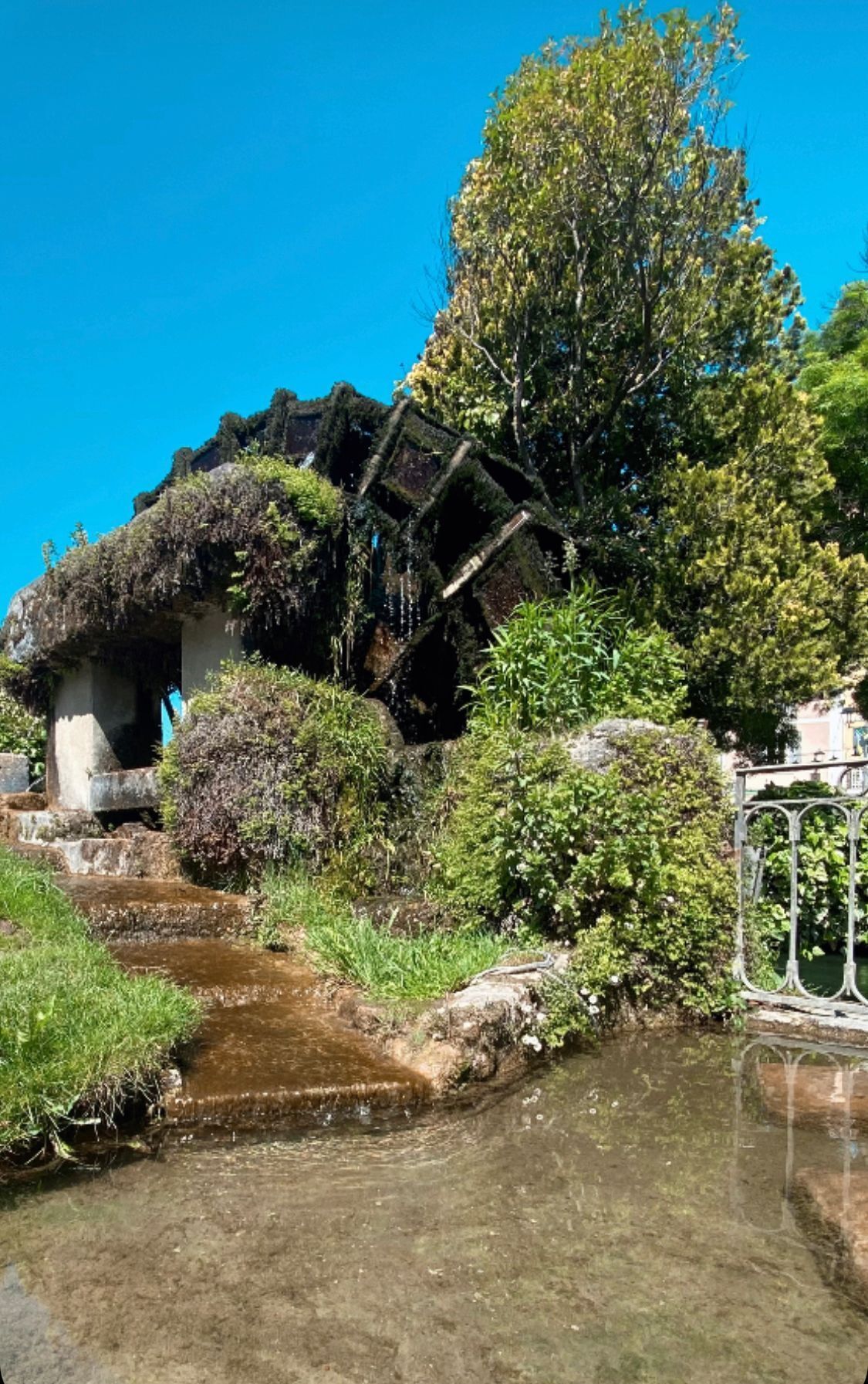 Een rustiek stenen gebouw met rieten dak, omgeven door weelderige begroeiing en een kleine beek.