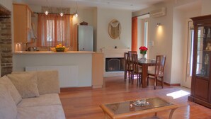A living room with a sofa, a glass coffee table, a dining area with chairs, a fireplace, and a kitchen with a refrigerator and fruit basket.