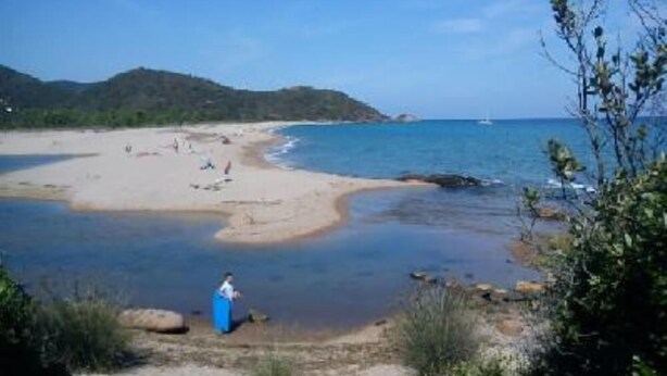 Ein Strand mit klarem, blauem Wasser, einem sandigen Ufer und einer Person in einem blauen Kleid am Wasser.