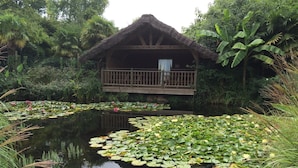 Une hutte au toit de chaume, dotée d’un balcon, entourée d’une végétation luxuriante et bordée d’un étang où flottent des nénuphars.