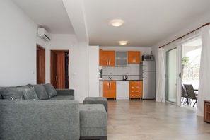 Uma sala moderna com um sofá e um banquinho cinza, uma cozinha com armários laranja e uma porta de vidro deslizante que dá para uma área externa.