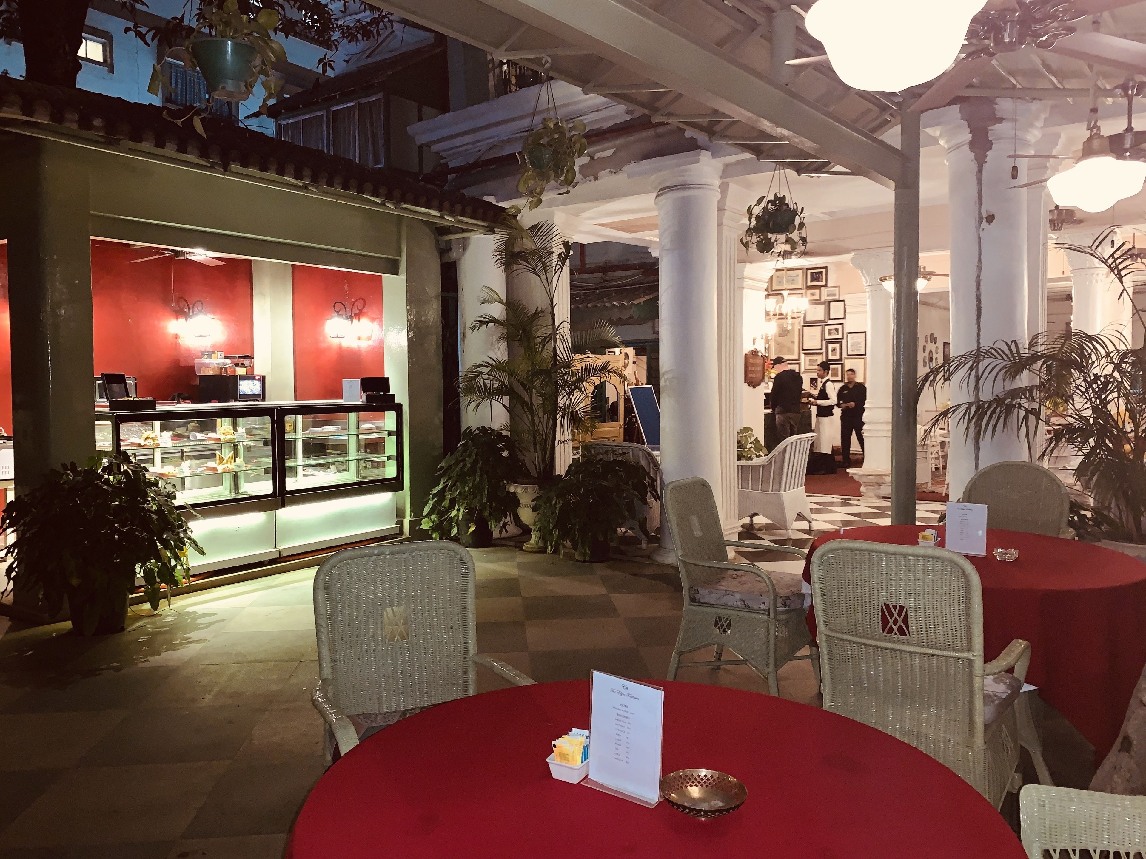 An outdoor cafe with wicker chairs and a red tablecloth, a dessert display case, and potted plants.