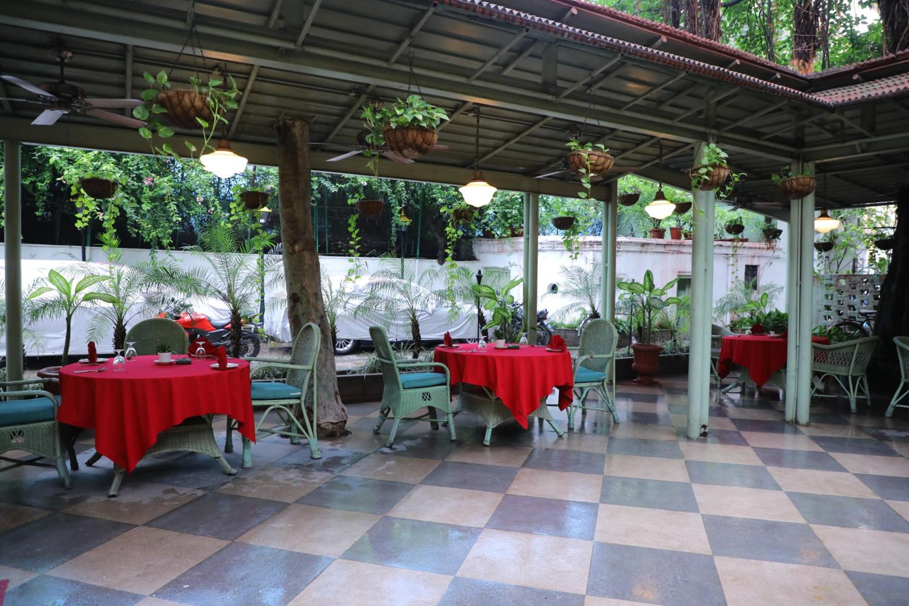 Un patio exterior con suelo a cuadros, sillas de mimbre verde, manteles rojos y macetas colgantes.