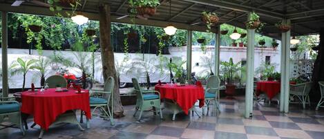 Un patio exterior con suelo a cuadros, sillas de mimbre verde, manteles rojos y macetas colgantes.