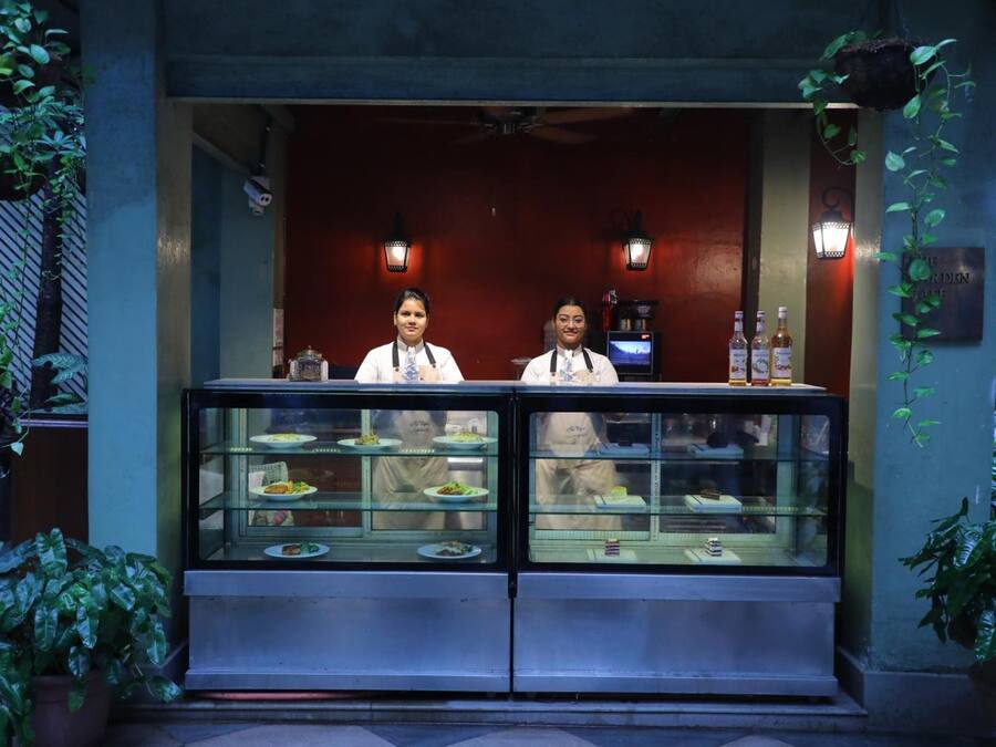 Un restaurante con un mostrador de vidrio lleno de comida, dos camareros detrás del mostrador y plantas decorativas alrededor de la entrada.