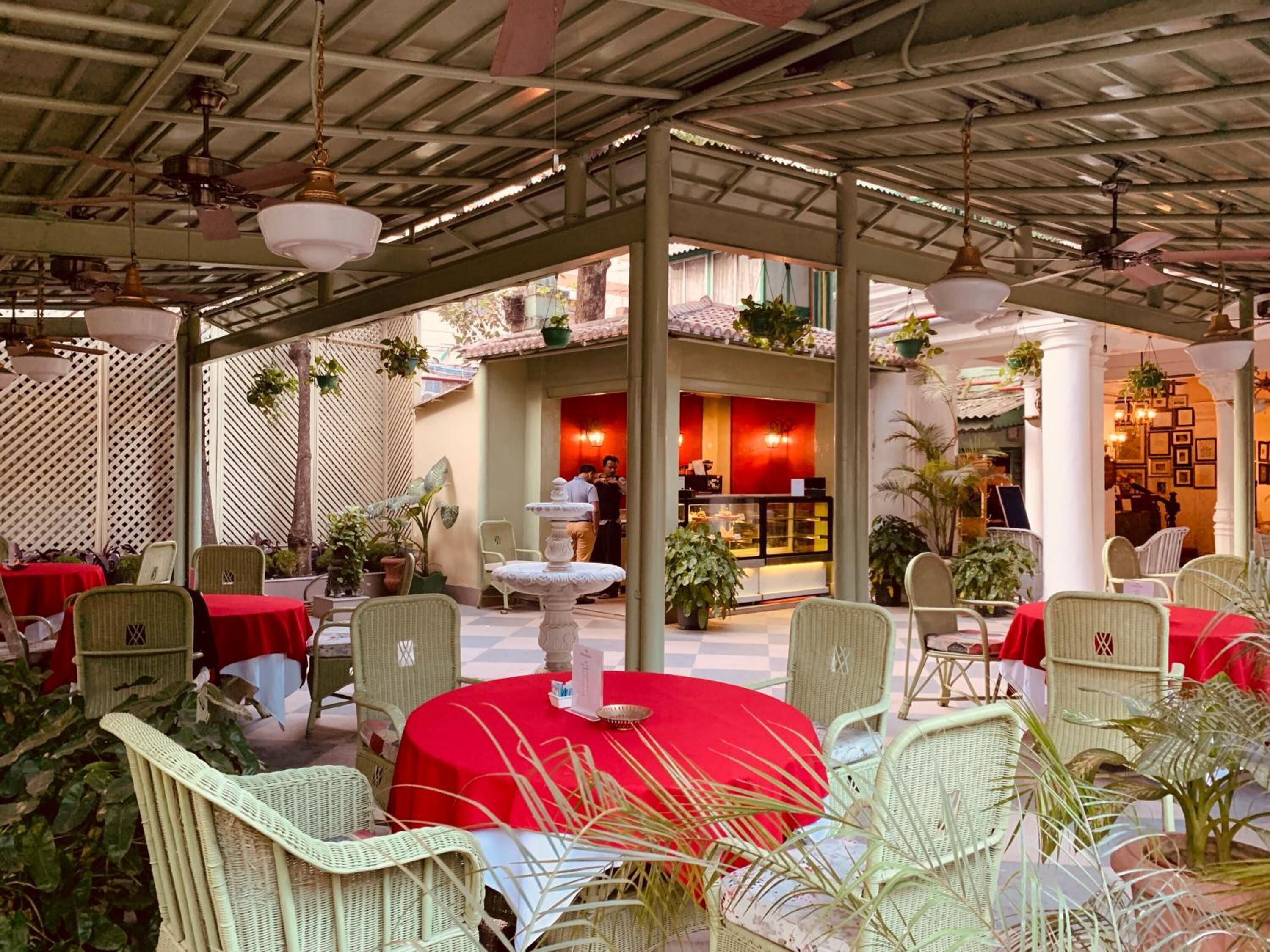 An outdoor dining area with wicker chairs and red tablecloths, a fountain, and a restaurant in the background.