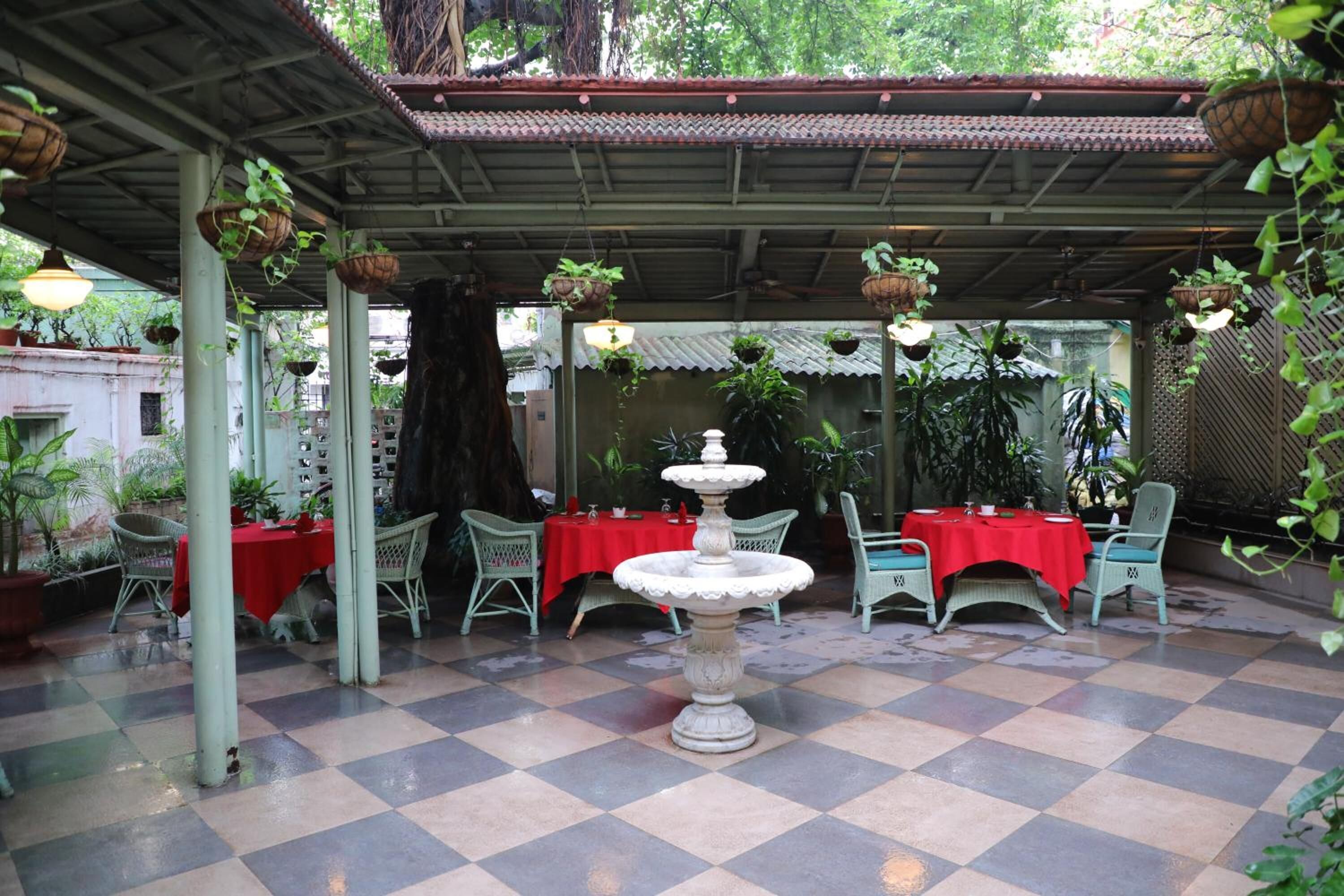 An outdoor patio with checkered flooring, red tablecloths, wicker chairs, hanging planters, and a central fountain.
