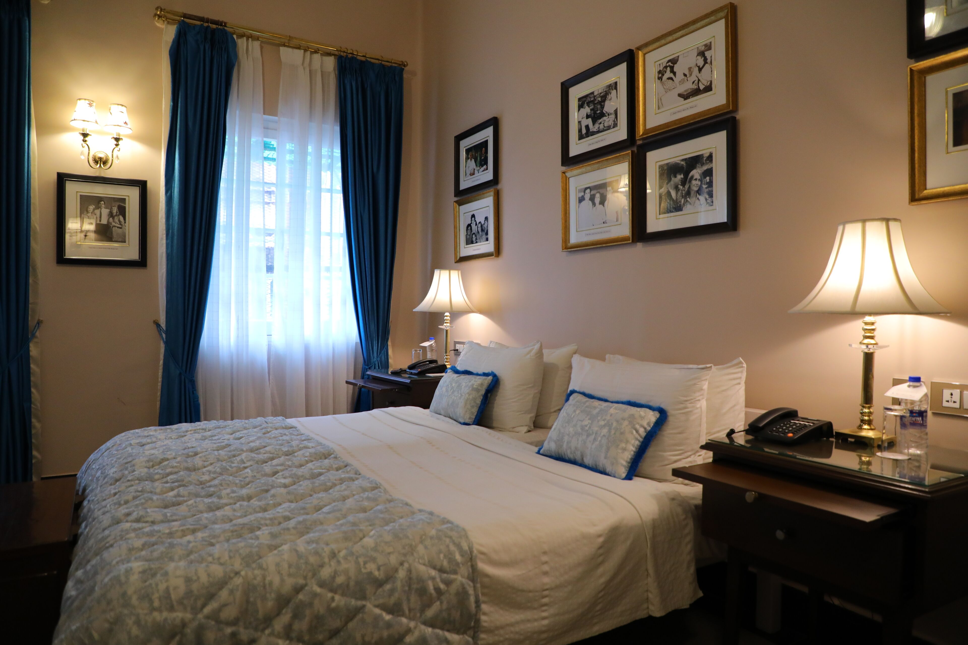 Habitación de hotel con una cama grande, dos mesitas de noche, un escritorio y una silla. En la pared hay cuadros enmarcados y una ventana con cortinas.