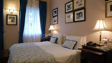 Habitación de hotel con una cama grande, dos mesitas de noche, un escritorio y una silla. En la pared hay cuadros enmarcados y una ventana con cortinas.