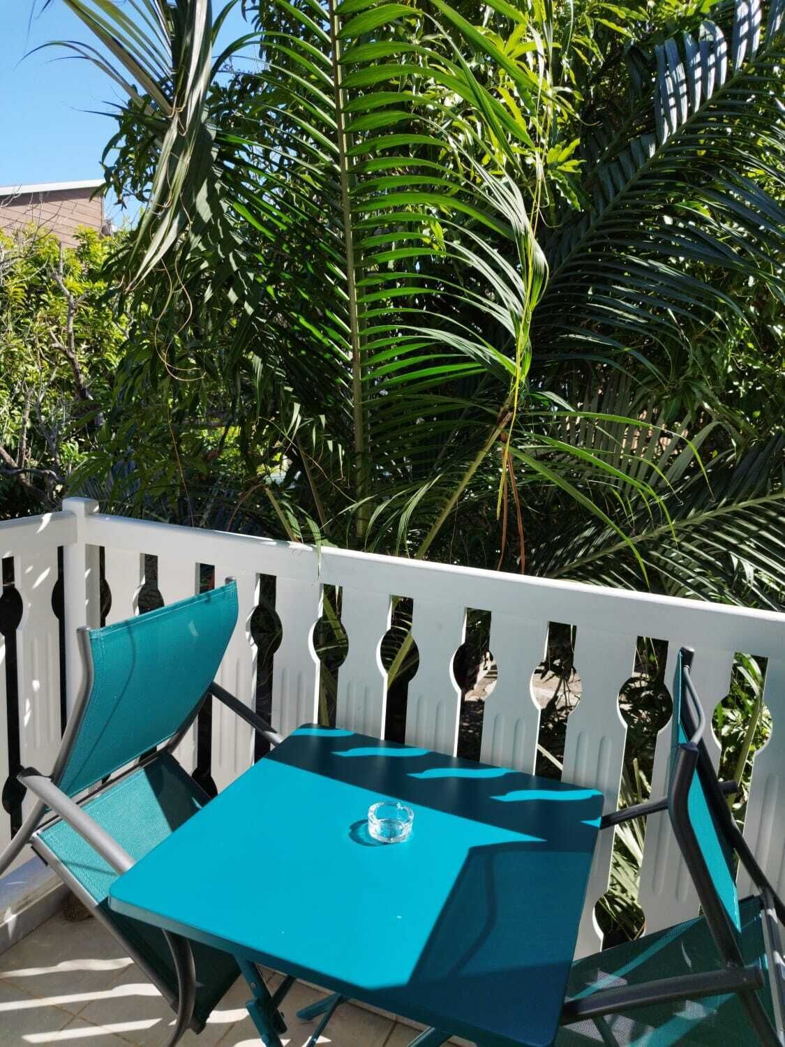 Een turquoise zitset met tafel en stoelen, geplaatst voor een witte hek, omgeven door weelderige begroeiing.