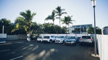 Uma área de estacionamento com vários carros, palmeiras e um edifício ao fundo.