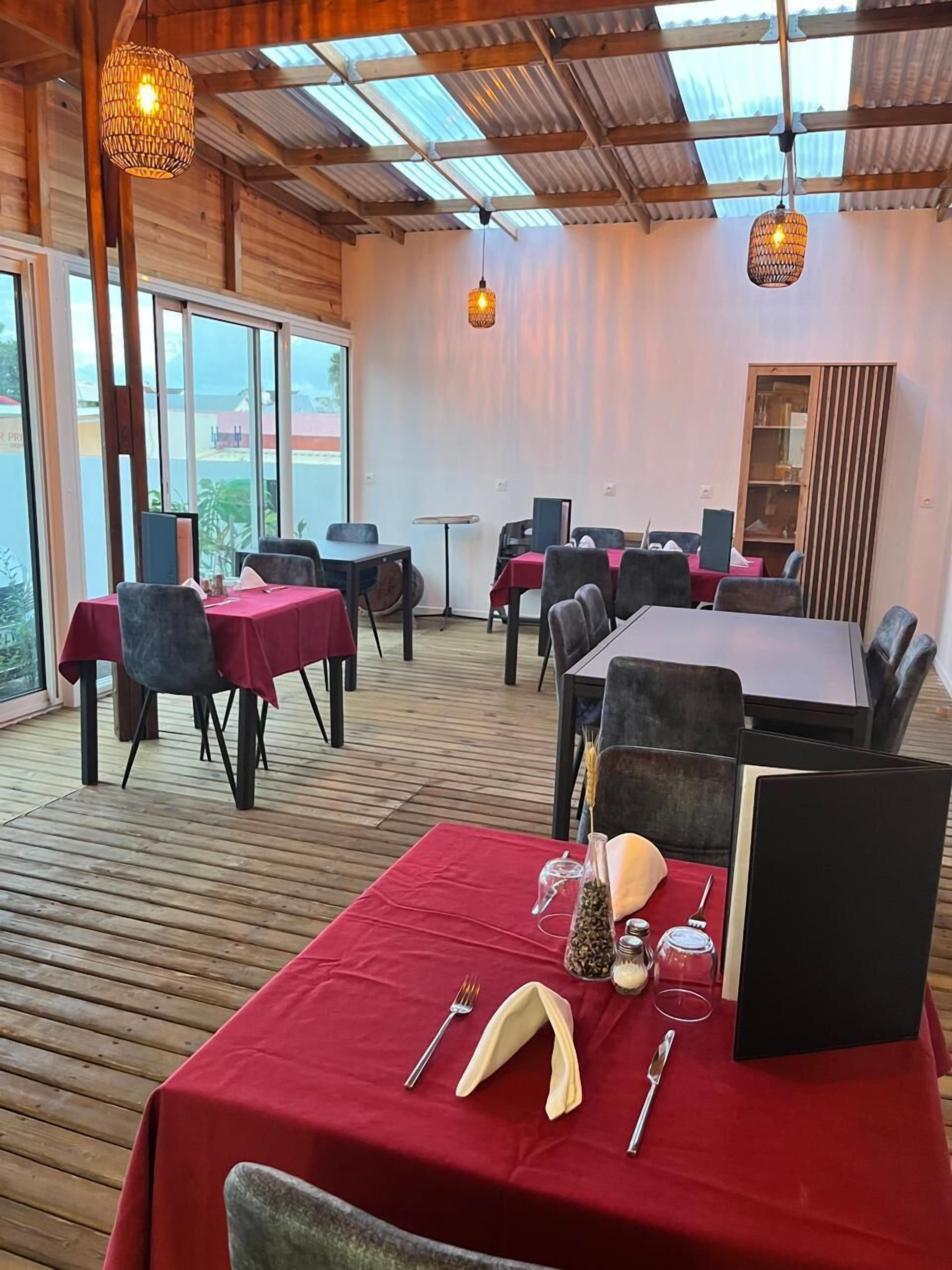 Ein Restaurant mit Holzboden, roten Tischdecken und schwarzen Stühlen. Hängelampen und ein großes Fenster bieten einen Blick nach draußen.
