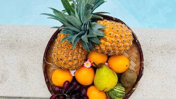 A fruit basket with pineapples, oranges, grapes, a pear, and a watermelon by a pool.