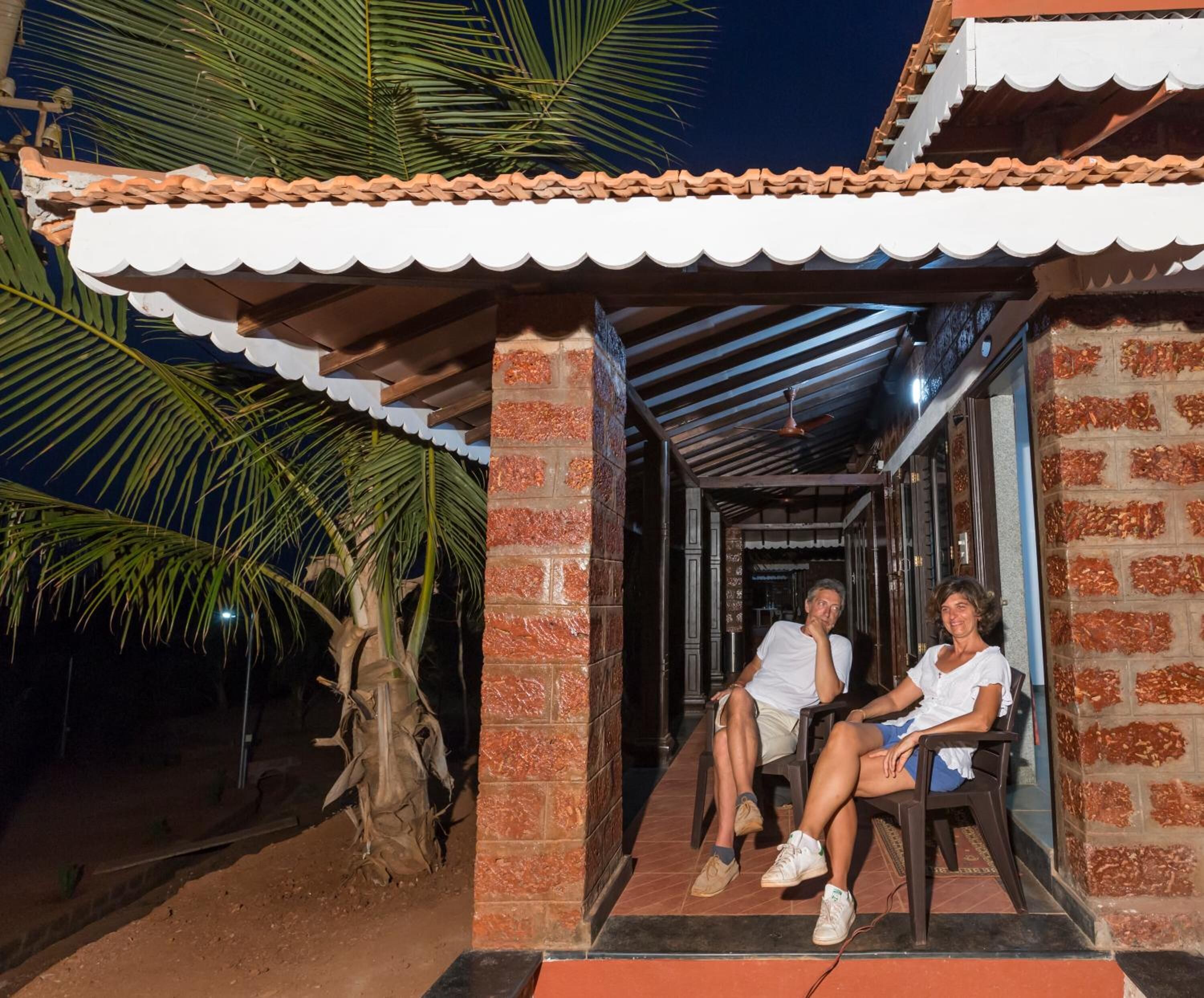 A covered outdoor seating area with two people seated, a palm tree, and a brick building with a tiled roof.