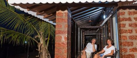 Un espacio de estar al aire libre cubierto con dos personas sentadas, una palmera y un edificio de ladrillo con tejado de tejas.