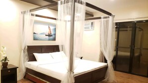 A four-poster bed with a canopy, a nightstand with a plant, a painting of a sailboat, and a sliding door. - A&A Garden Suites (Amadeo)