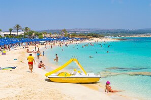 Une plage bondée, avec une eau turquoise cristalline, un bateau jaune et des gens qui profitent du soleil.