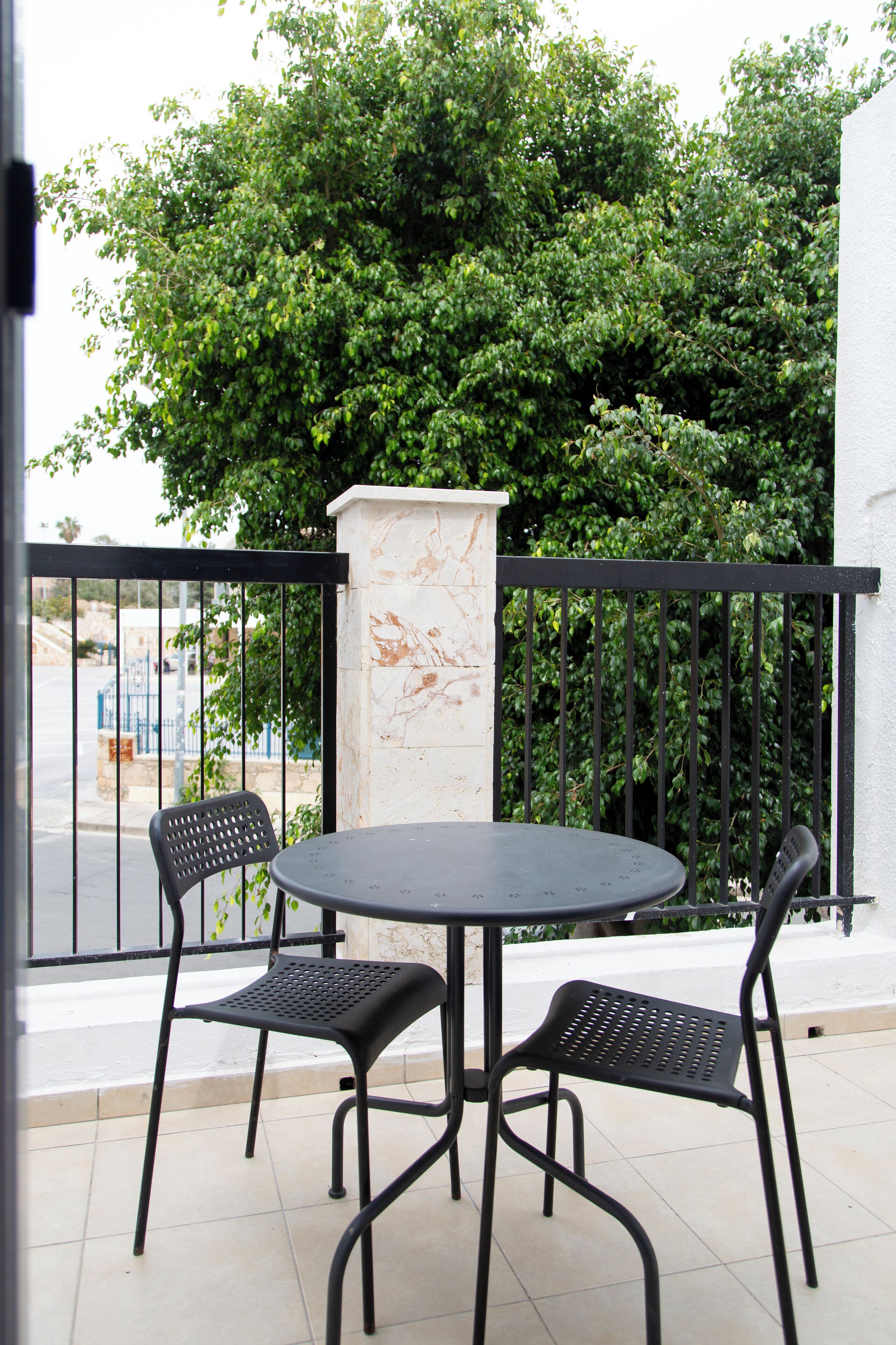 A round table with two chairs on a balcony with a black railing, a white pillar, and a view of a street and greenery.