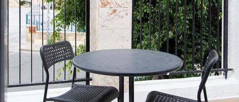 A round table with two chairs on a balcony with a black railing, a white pillar, and a view of a street and greenery.