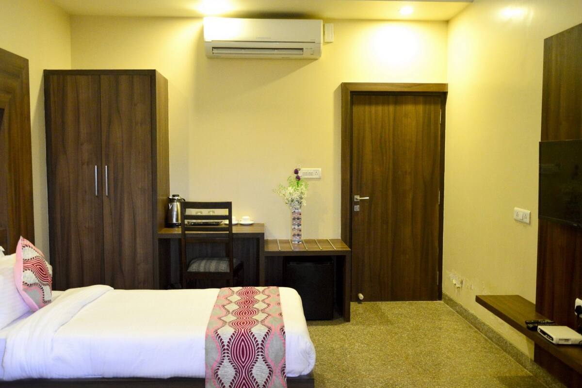 A hotel room with a bed, a desk, a chair, a small table with a vase, and a TV.