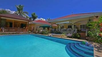 A swimming pool with a curved edge, surrounded by a house with a red roof and a covered patio area.