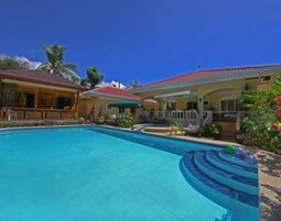 Una piscina de borde curvo, rodeada por una casa con techo rojo y un patio cubierto.