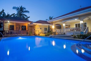Una piscina con zona cubierta, edificio de fachada de piedra y una palmera al fondo.