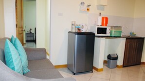A compact living space with a grey sofa, a small kitchen area including a microwave and a refrigerator, and a trash bin. - Alona Austria Resort (Panglao)