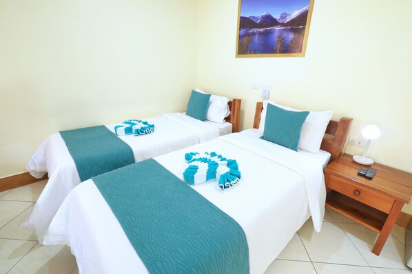 Habitación de hotel con dos camas, cada una con cubrecama color aguamarina y ropa de cama blanca, una mesita de noche de madera con lámpara, y un cuadro enmarcado en la pared.