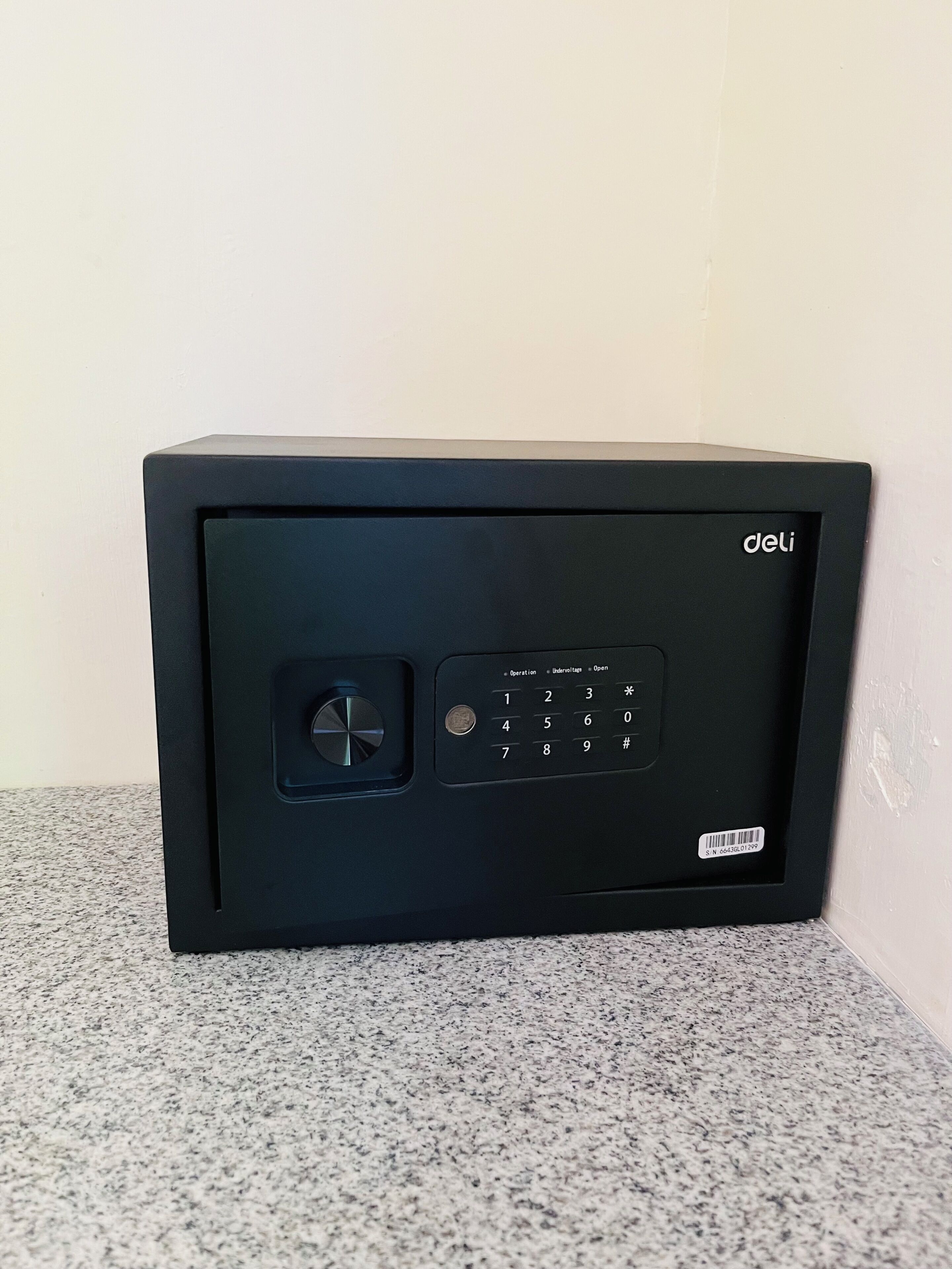 A black safe with a keypad and a dial lock, placed on a speckled floor against a plain wall.