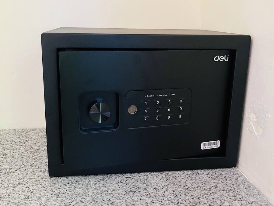 A black safe with a keypad and a dial lock, placed on a speckled floor against a plain wall.
