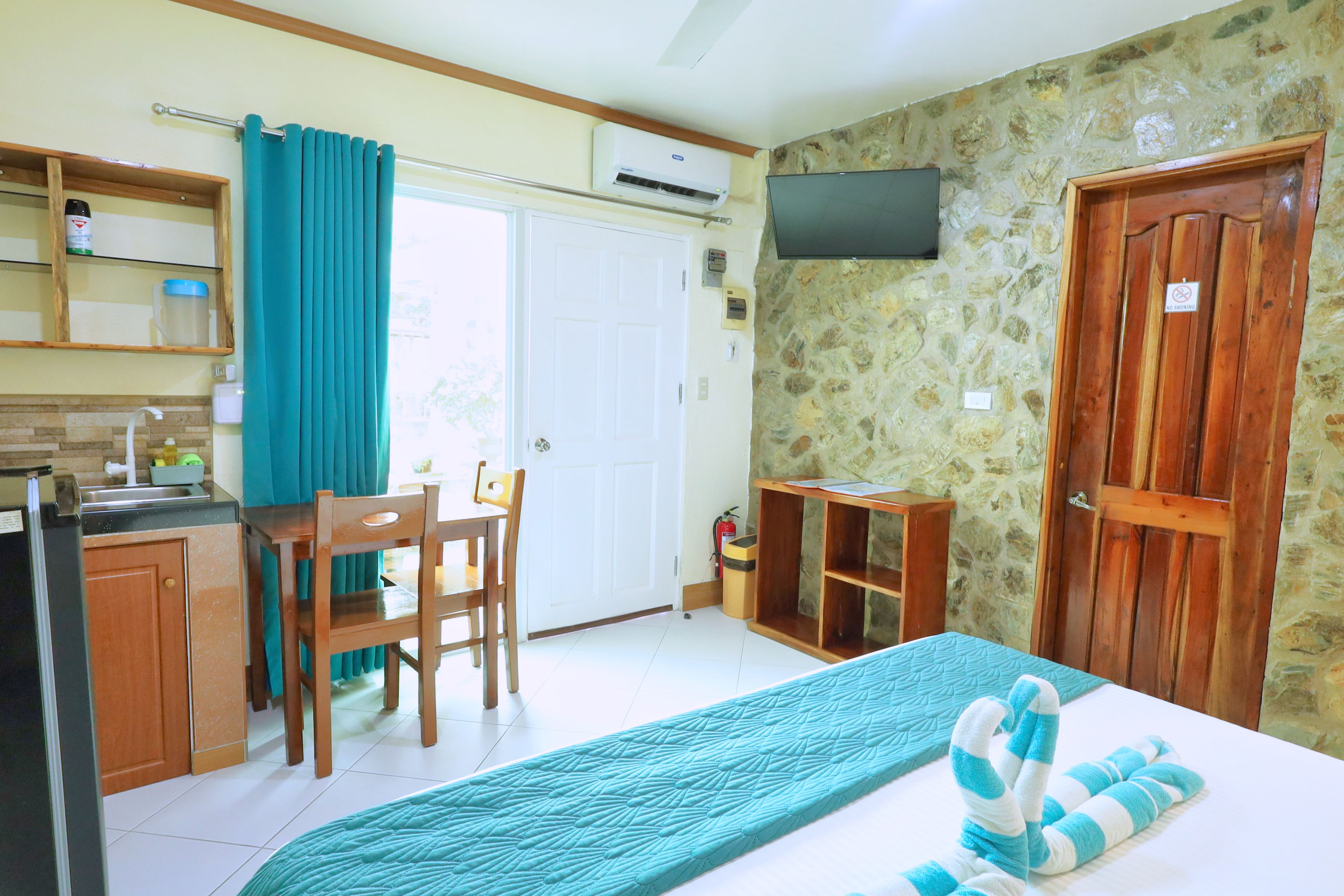A room with a bed, a desk, a chair, a television, and a kitchen area.