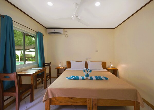 A hotel room with a bed, two bedside tables, a desk, and chairs. There is a window with curtains, an air conditioning unit, and a ceiling fan.