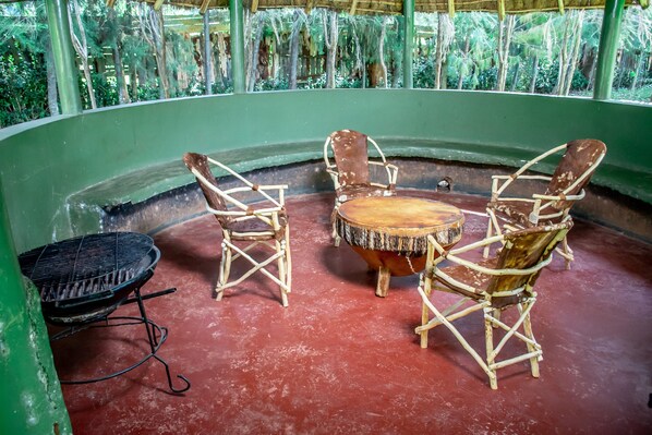 A covered outdoor seating area with a round table and four wicker chairs, a grill, and a green wall.