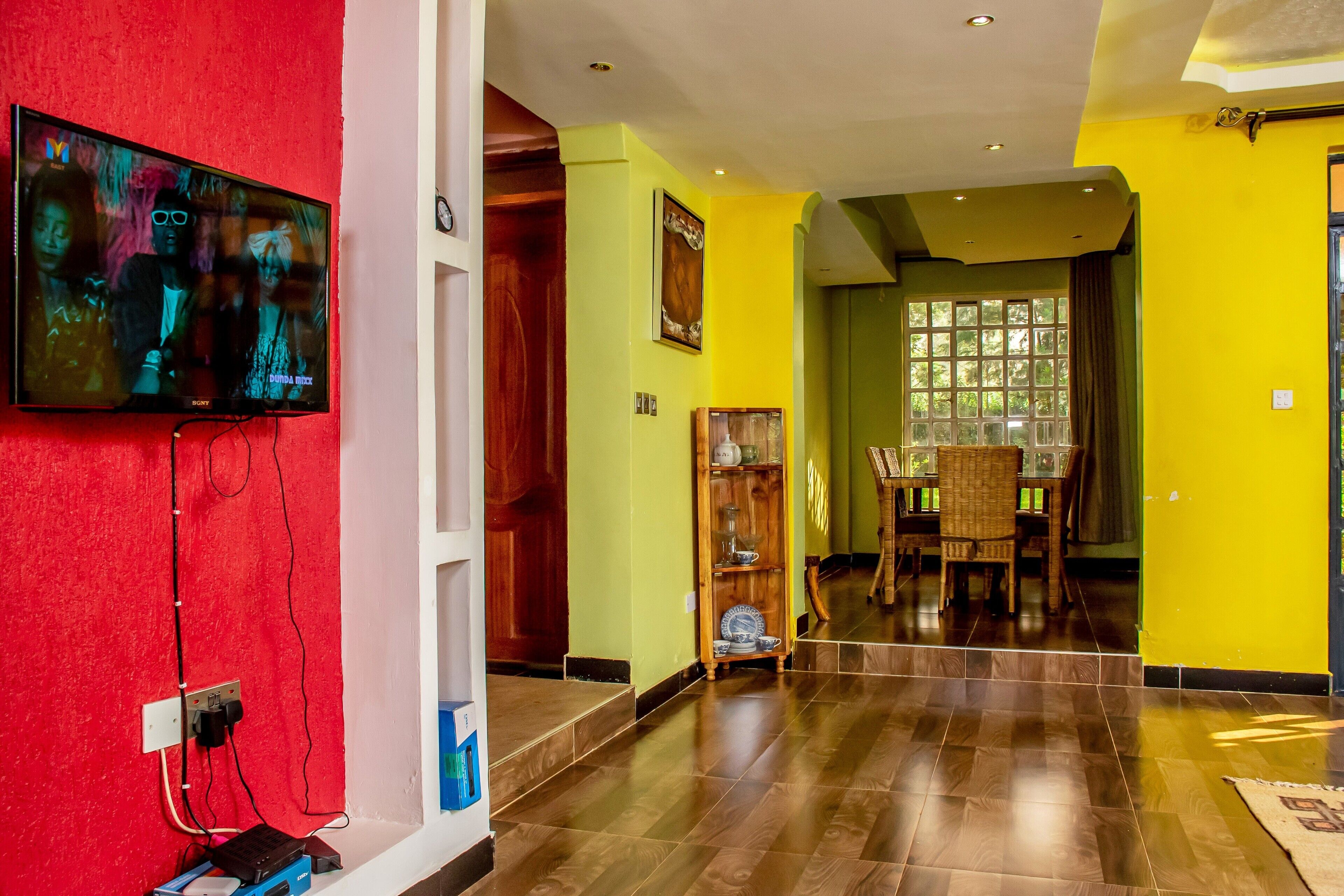 Un pasillo con un televisor montado en una pared roja, suelo de madera y una zona de comedor con mesa y sillas.