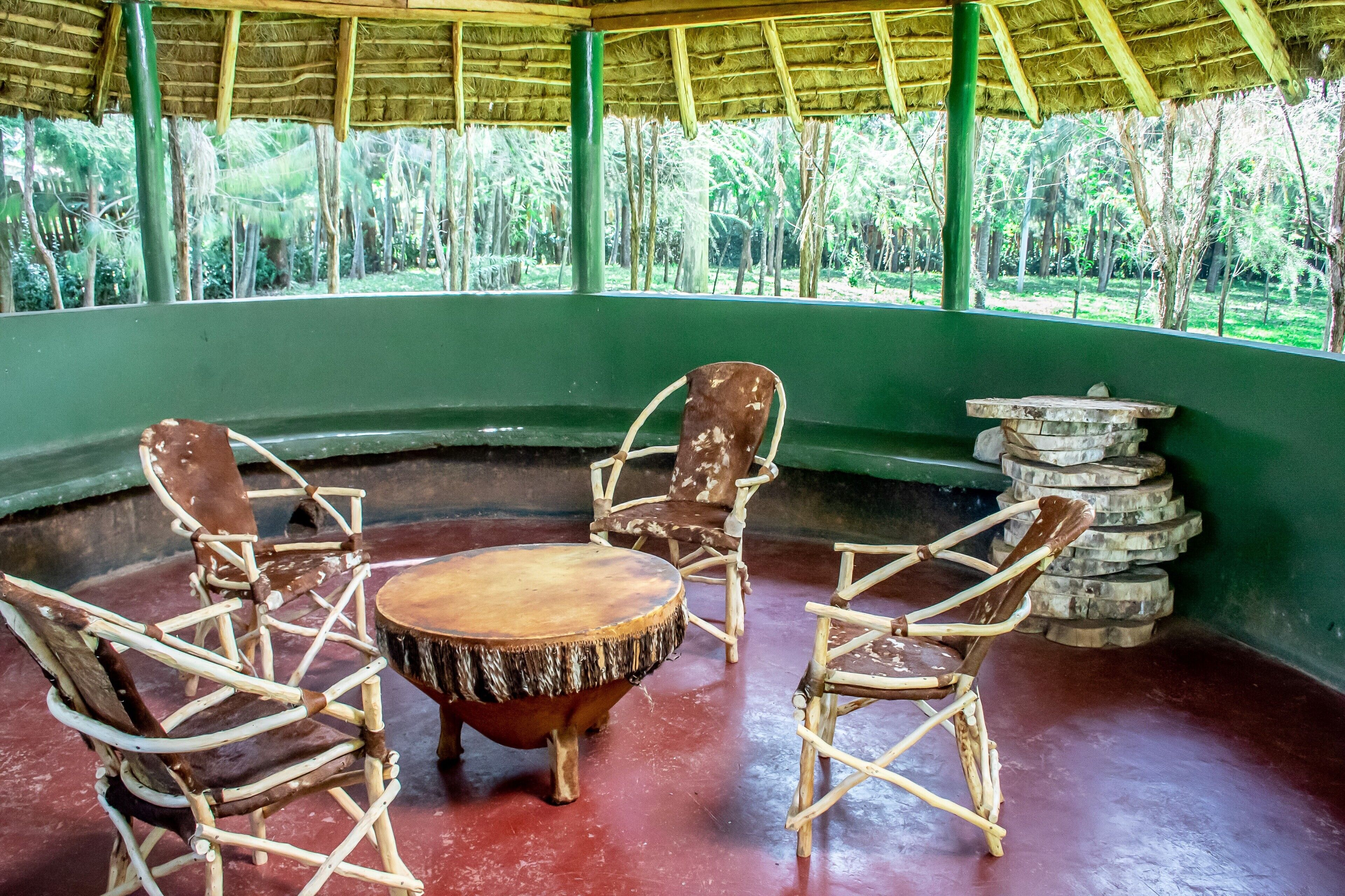 A covered seating area with a round wooden table and four wicker chairs, surrounded by a green wall and a view of a tropical forest.