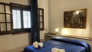 A room with a bed, a window with blinds, a painting on the wall, and a bedside lamp.