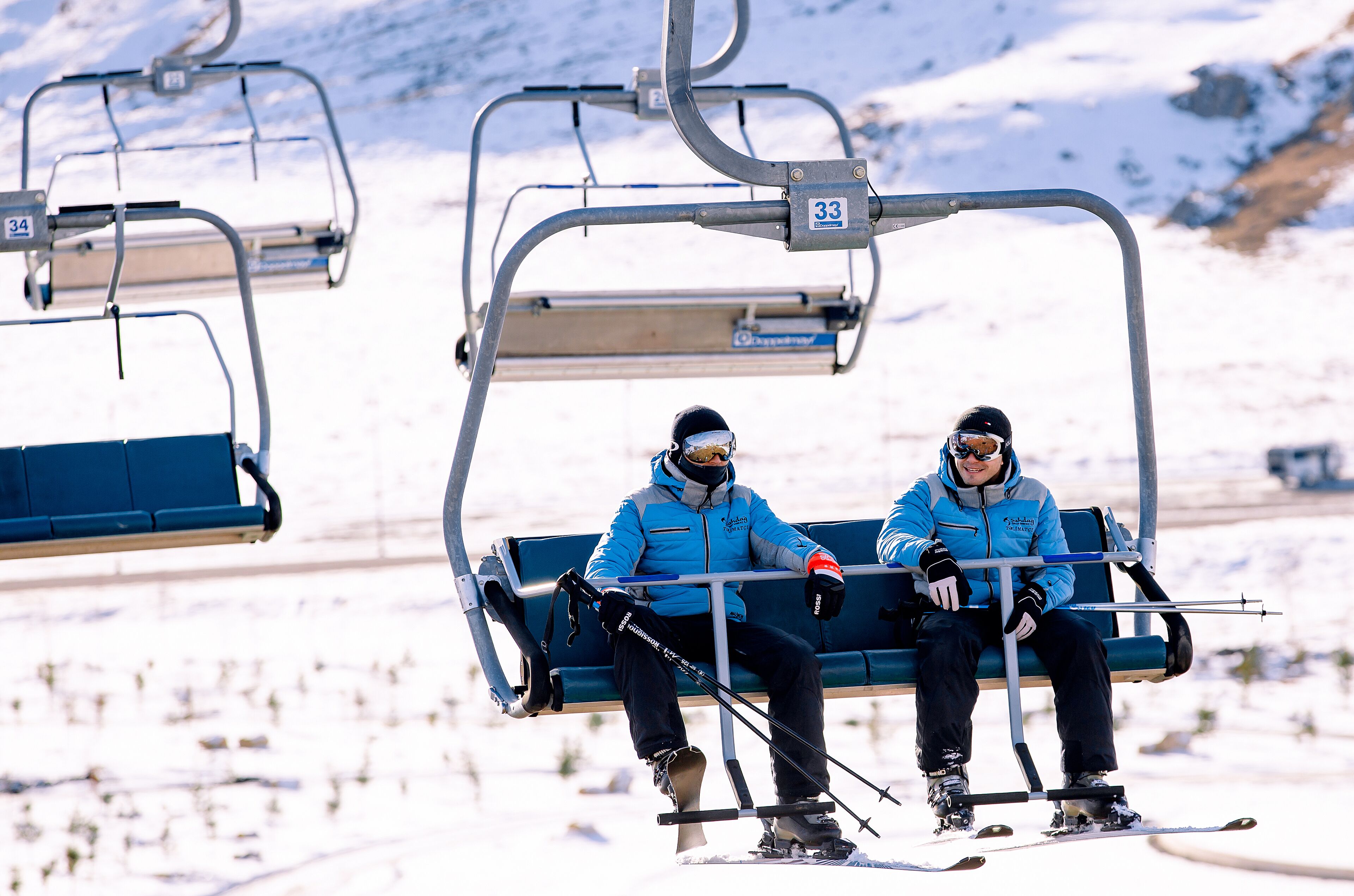 Two people sitting on a ski lift chair with ski poles, wearing blue jackets and black pants.