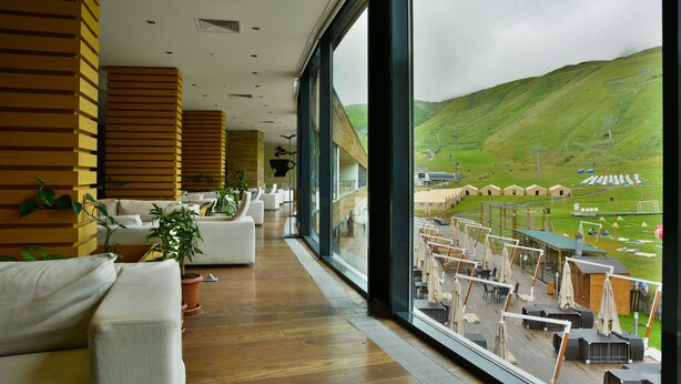 Un intérieur d’hôtel moderne avec un plancher en bois, des canapés blancs et de grandes fenêtres offrant une vue sur une colline verdoyante et un espace de détente extérieur.