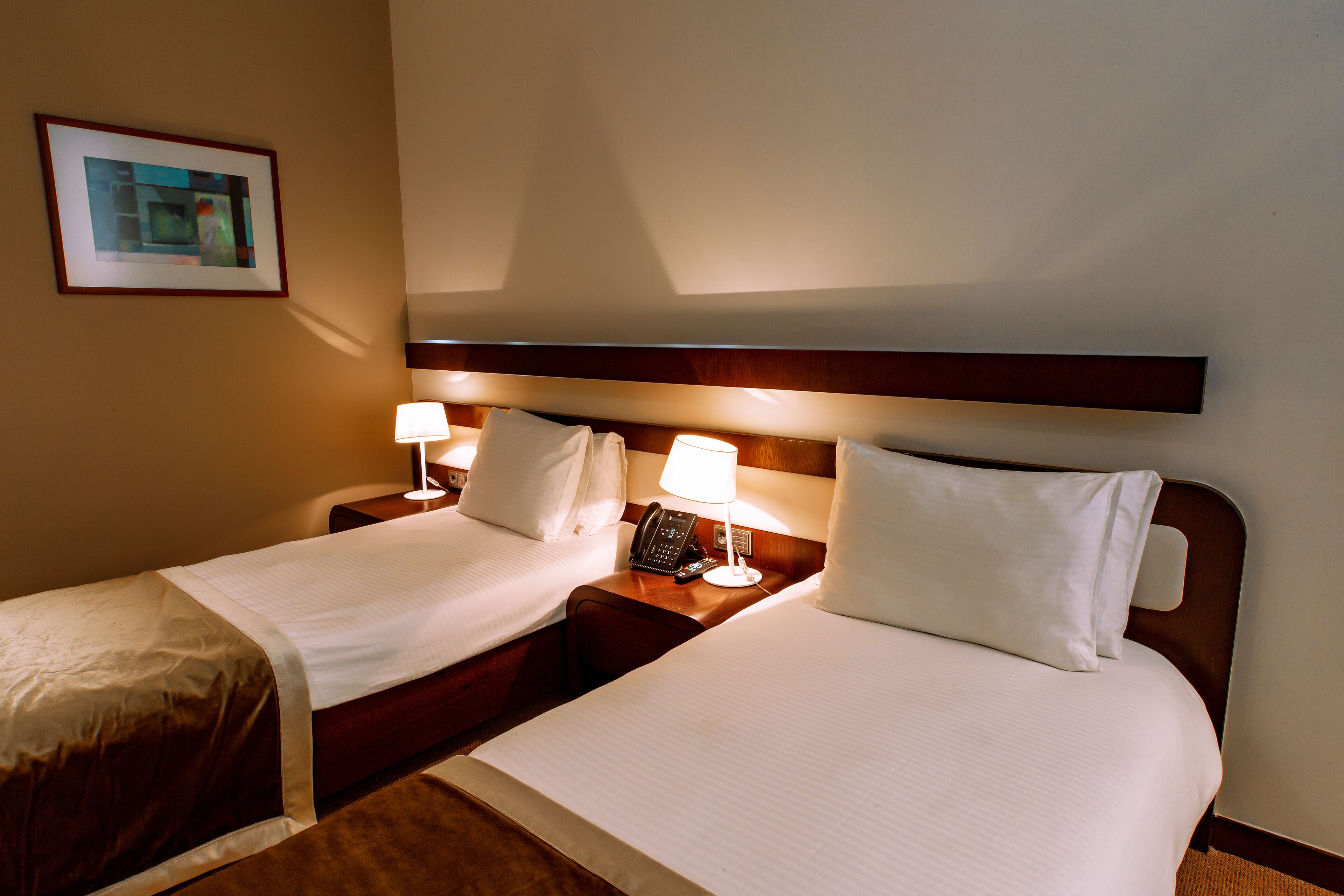 Een hotelkamer met twee bedden, nachtlampjes, een telefoon en een ingelijst schilderij aan de muur.