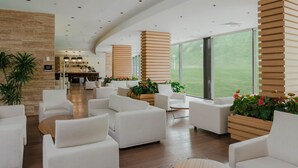 Un hall d’entrée spacieux, doté de canapés blancs, d’éléments décoratifs en bois et de grandes fenêtres offrant une vue sur l’extérieur.