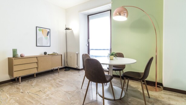 Un comedor con una mesa redonda, dos sillas, una lámpara de pie, un armario y un cuadro en la pared.
