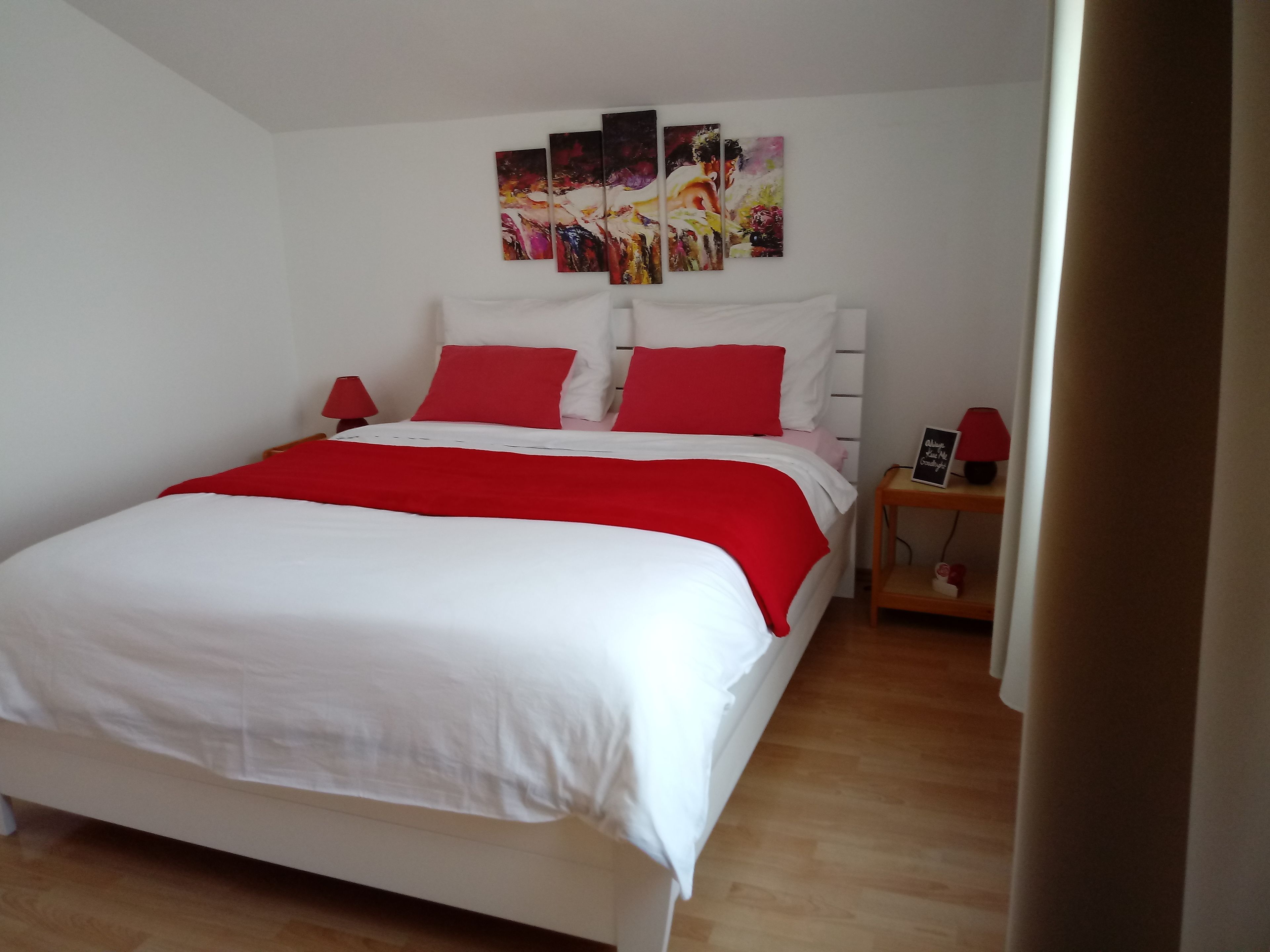A neatly made bed with white and red bedding, a bedside table with a lamp, and a wall with three abstract paintings.